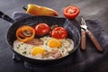 Fried eggs with bacon and tomatoes on an old cast iron pan and cutlery on a gray table. Royalty Free Stock Photo