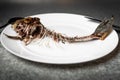 Fried fish skeleton, spine and bones on a white plate with a black knife and fork on a gray background Royalty Free Stock Photo