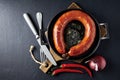 Fried sausage rolled into a ring on a cast iron pan with cutlery, a wooden cutting board, hot peppers and red onions on Royalty Free Stock Photo