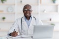 Friendly african american male doctor working with laptop in modern office, taking notes, planning his day Royalty Free Stock Photo