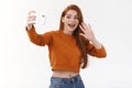 Friendly attractive redhead modern woman contact friend abroad using internet, holding smartphone horizontally waving at