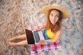 A friendly attractive young Caucasian woman in a straw hat with a laptop in her arms sits on the sandy beach and looks Royalty Free Stock Photo