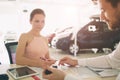 Friendly car salesman talking to a young woman and showing a new car inside showroom Signing of contract.