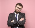 Friendly Caucasian bearded man in glasses and a suit stands with crossed arms and smiles cutely. Pink background.