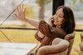 Friendly charming brunette woman with long curly hair sitting at the window in cafe with mobile phone and waving to