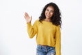 Friendly cute african-american curly-haired girl in yellow sweater raise palm and waving, saying hello, greeting guests