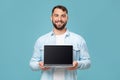 Friendly glad adult european man showing laptop with blank screen, isolated on blue background, free space