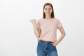 Friendly-looking confident young happy girl in casual clothes, holding hand in pocket while pointing left with thumb and Royalty Free Stock Photo