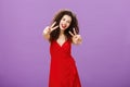 Friendly-looking peaceful european female with curly haircut in elegant red dress showing peace or victory signs
