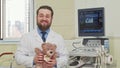 Friendly male pediatrician holding teddy bear toy, waving to the camera