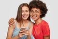 Friendly mixed race women embrace and smile together, watch funny video content on cellular, pose against white Royalty Free Stock Photo