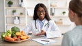 Friendly nutritionist black woman giving consultation to patient and demonstrating plate with fresh fruit and vegetables