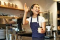 Friendly smiling barita girl waving at client, holding their cup of coffee, prepared takeaway order, working in cafe Royalty Free Stock Photo