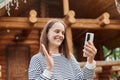 Friendly smiling young beautiful caucasian woman wearing striped shirt using smartphone standing near wooden house waving hand Royalty Free Stock Photo