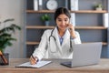 Friendly smiling young cute indian female therapist in uniform talking by phone with patient in office interior Royalty Free Stock Photo