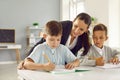 Friendly teacher helps small schoolchildren with test tasks in elementary school classroom.