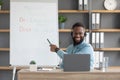 Friendly young african american man tutor showing rules of english at blackboard and looks at pc