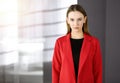 Friendly young business woman or female student dressed in red coat is standing straight and looking at camera in a Royalty Free Stock Photo