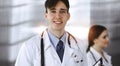 Friendly young male doctor standing with clipboard in modern clinic. Female colleague is busy with filling up of