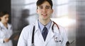 Friendly young male doctor standing with clipboard in modern clinic. Female colleague is busy with filling up of