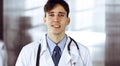 Friendly young male doctor standing with clipboard in modern clinic. Female colleague is busy with filling up of