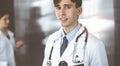 Friendly young male doctor standing with clipboard in modern clinic. Female colleague is busy with filling up of