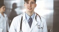 Friendly young male doctor standing with clipboard in modern clinic. Female colleague is busy with filling up of