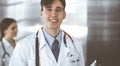 Friendly young male doctor standing with clipboard in modern clinic. Female colleague is busy with filling up of