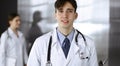 Friendly young male doctor standing with clipboard in modern clinic. Female colleague is busy with filling up of