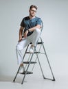 Friendly young male workman repairman in uniform, doctor sitting on stepladder. Confident guy on ladder, studio portrait