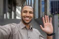 Friendly young man in casual attire waving hello in urban setting, expressing joy and approachability Royalty Free Stock Photo
