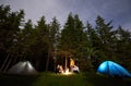 Friends around a campfire between tents amid powerful firs under a starry sky. Nightcaping in the forest.