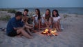 Friends sit around bonfire, drink beer, sing to guitar, fry sausages on sandy beach. Young mixed race group of men and