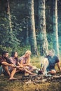 Friends talking around campfire. Men in lumberjack shirts cooking sausages outdoors. Two couples camping in woods