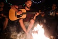 Friends travelers chilling at big bonfire, singing songs and playing guitar at camp in the night forest. Group of people resting Royalty Free Stock Photo