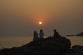 Friends, two girls and a guy sit on a rock against the backdrop of the ocean and purple pink gray