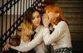 A friendship between two schoolgirls in uniform sitting on a staircase. A ginger Asian girl is whispering a secret into the ear of