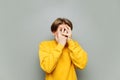 Frightened young man in yellow clothes stands on a gray background, hiding with his hands on his face and peeping into the cameo. Royalty Free Stock Photo