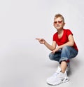 Frolic blond kid boy in red t-shirt, blue jeans, sneakers and sunglasses sits squatted pointing finger at copy space