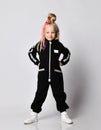 Frolic blonde kid baby girl with braids in black jumpsuit and sneakers stands with hands on waist and makes grimaces