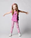 Frolic blonde kid girl in summer clothing colorful pants and t-shirt stands with her arms outstretched and screams