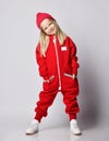 Playful blonde kid girl in white sneakers, red overall jumpsuit and winter hat stands swaying holding hands in pockets