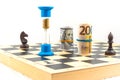 Front angle view of chess board with two black chess pieces, an hourglass with falling sand within and rolled euro bank