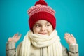 Front close-up portrait of little girl in red winter hat and white scarf on blue background, raised her hands up, looks Royalty Free Stock Photo