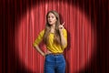 Front crop view of beautiful young girl in yellow jumper and blue jeans, standing in spotlight against red stage curtain Royalty Free Stock Photo