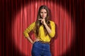 Front crop view of pretty woman standing in spotlight at red stage curtain, one hand on hip, other hand doing silencing Royalty Free Stock Photo