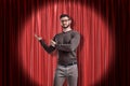 Front crop view of young handsome man in casual clothes and glasses standing in spotlight against red stage curtain and