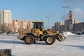 A front-end wheel loader removes snow from a city square after a snowfall against the backdrop of residential buildings on a sunny