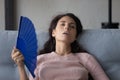 Stressed millennial woman waving paper fan at home.