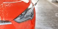 Front headlight of red car washed in self serve carwash, high pressure water stream spraying on glass